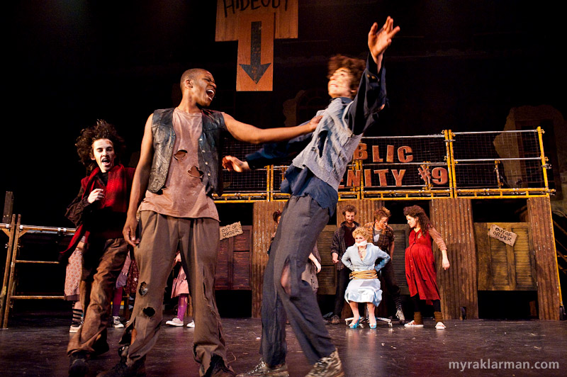 Pioneer Theatre Guild: Urinetown | Hot Blades Harry (John Spalding), center, shows Tiny Tom (Russel Norris) the meaning of “Urinetown” in What is Urinetown? I love the enormous sign (partially visible in the photo) that reads “Secret Hideout” — and even features a helpful arrow showing just where it is. This is typical of the self-referential (and in this instance, self-negating) humor of the show.