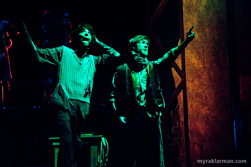 Pioneer Theatre Guild: Urinetown | NEW PHOTO: Slick “flashback” interludes…
Old Man Strong (right): Bobby! Bobby, reason with the woman. I’m a little short this morning.
Tiny Tom (left, tossing silver glitter): No shorter than yesterday. Unless I’ve grown.